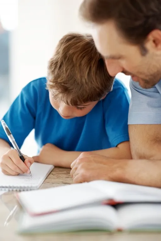 Dziecko przy stole piszące w zeszycie oraz siedzący obok mężczyzna patrzący na zeszyt